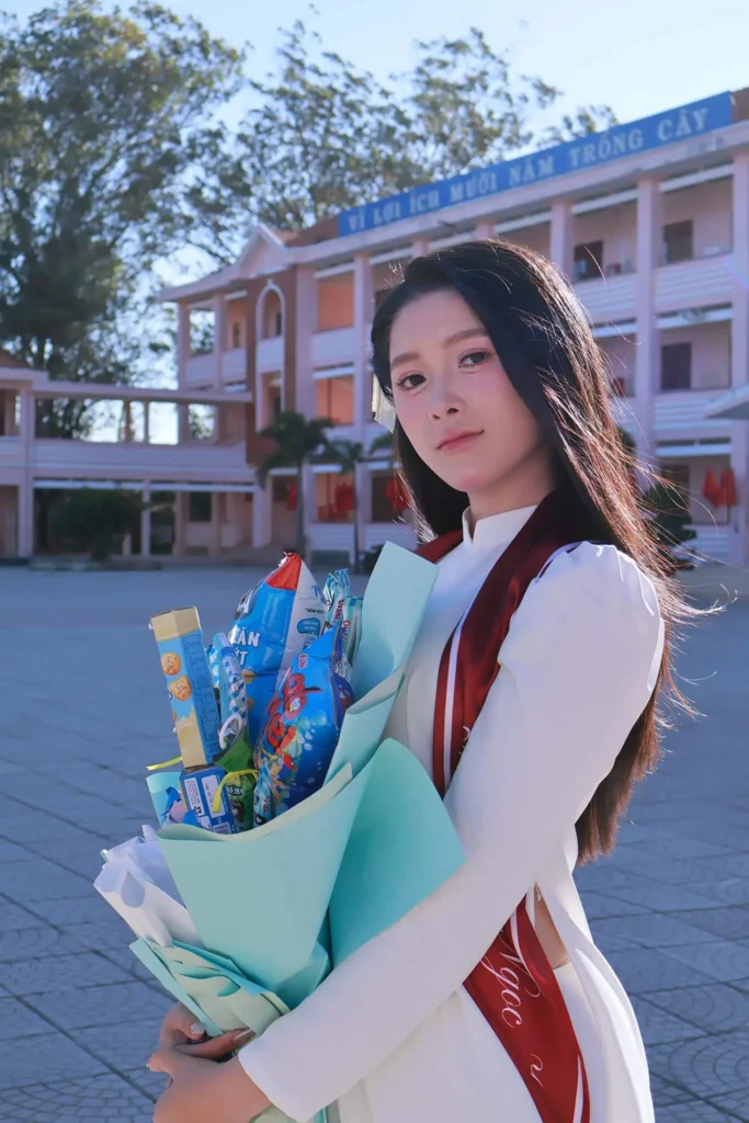 Nữ sinh mặc áo dài trắng ôm bó hoa đứng trước dãy phòng học trong buổi chụp kỷ yếu