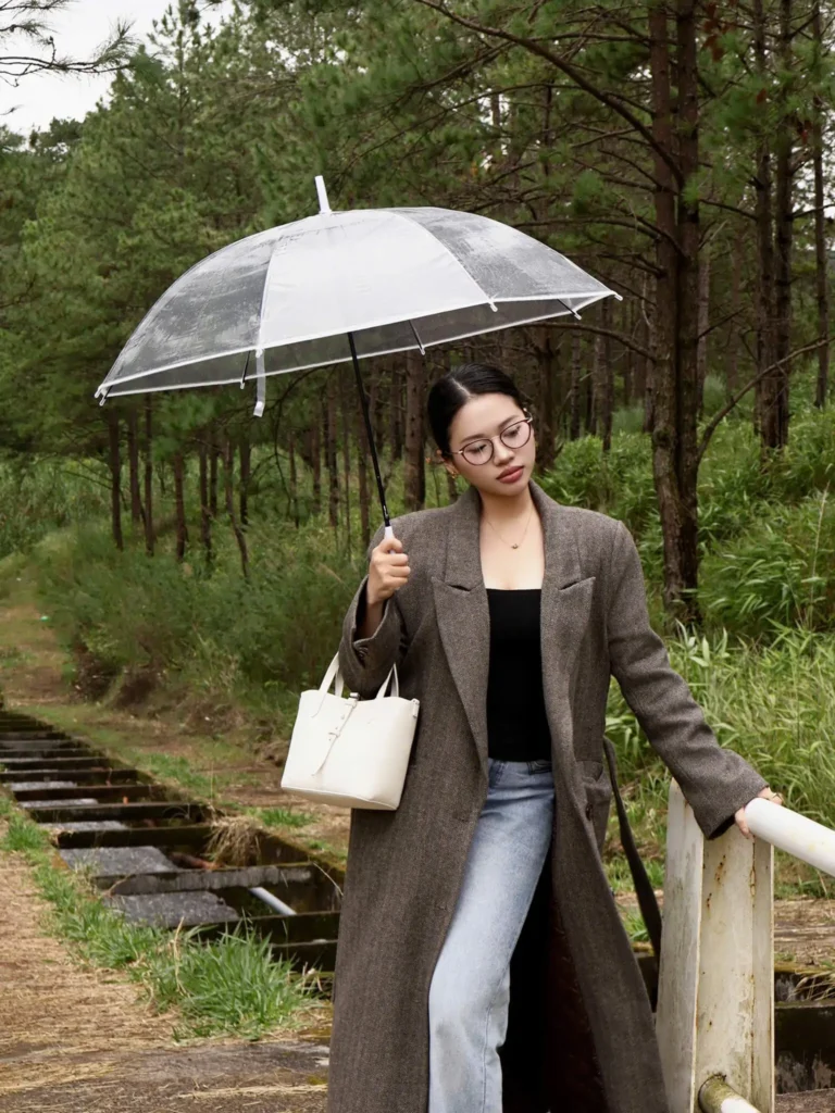 Cô gái mặc áo khoác dài cầm ô trong suốt đứng bên lối đi giữa rừng thông Đà Lạt lúc trời mưa nhẹ