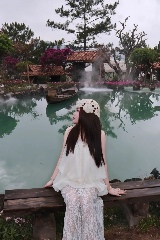 Cô gái mặc váy trắng ngồi trên lan can gỗ nhìn ra mặt hồ phủ sương, phía sau là dãy nhà mái ngói trong khu vườn ở Đà Lạt