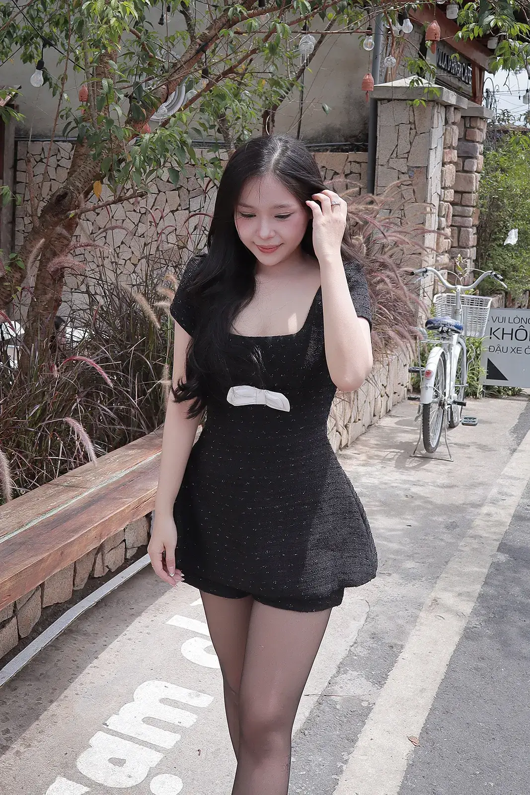 Cô gái mặc váy đen đi trên lối đi giữa bụi cỏ lau, phía sau có xe đạp và tường đá