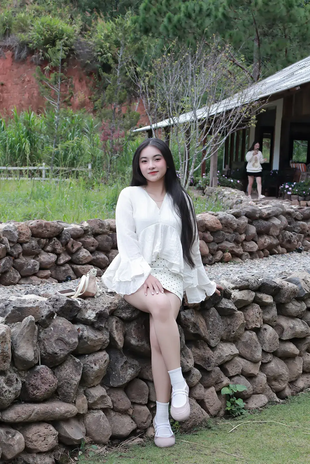 Cô gái mặc váy trắng ngồi trên bờ kè đá, phía sau là dãy nhà mái tôn và sườn đồi đất đỏ