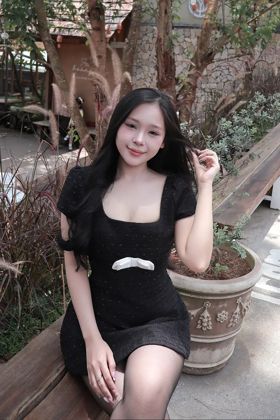 Cô gái mặc váy đen ngồi trên ghế gỗ giữa chậu cây và bụi cỏ lau trong khuôn viên quán