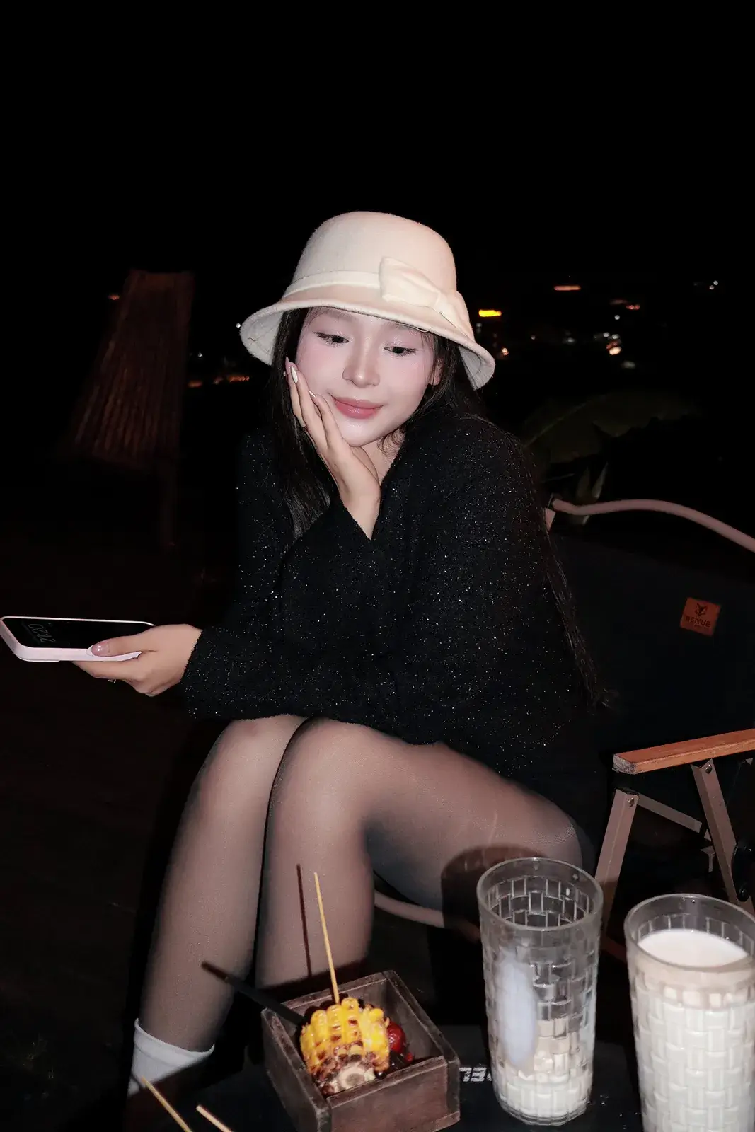 Cô gái đội mũ trắng ngồi ở bàn ngoài trời ban đêm cùng đồ uống, phía xa là ánh đèn thành phố