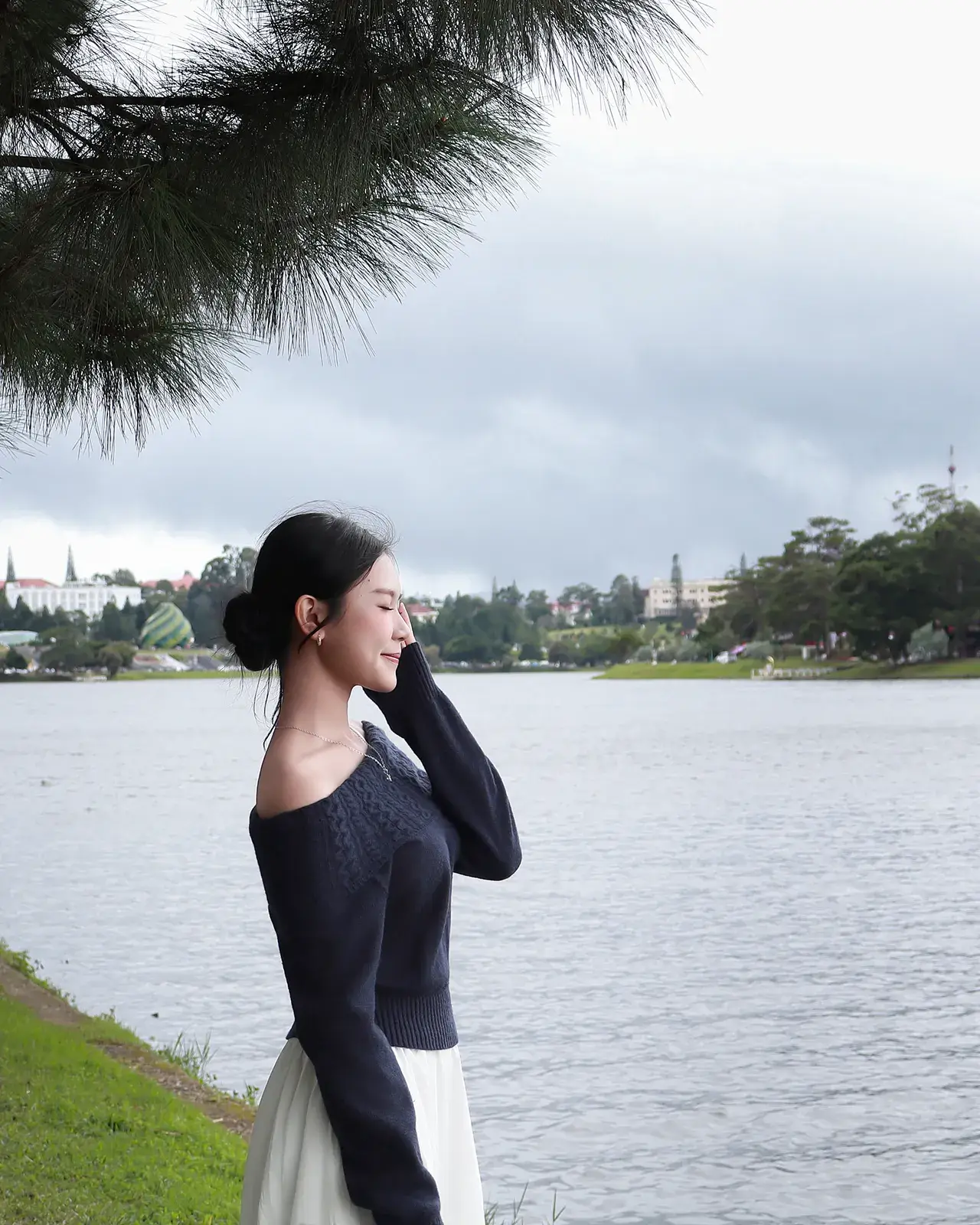 Cô gái đứng bên hồ dưới tán thông, bờ bên kia là khu phố và đồi cây