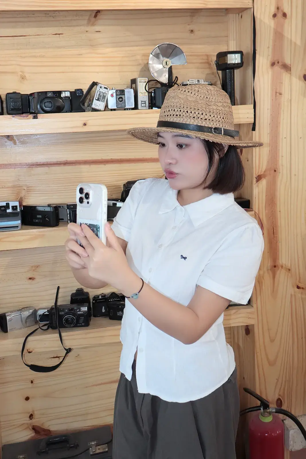 Cô gái đội mũ cói cầm điện thoại đứng trước kệ gỗ trưng bày nhiều máy ảnh film