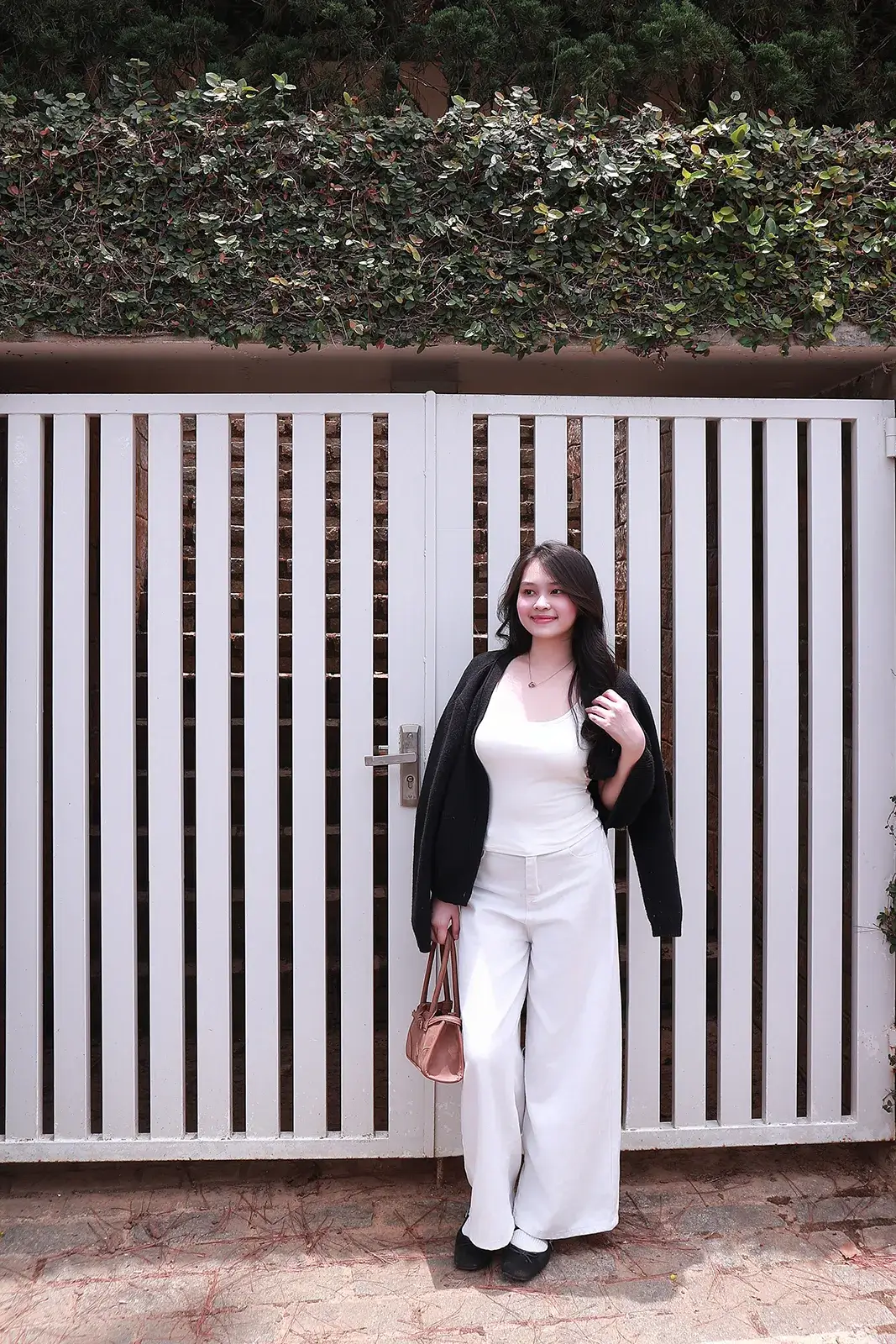 Cô gái mặc áo trắng khoác cardigan đen và quần ống rộng, đứng trước cổng rào gỗ sơn trắng có hàng cây xanh phía trên