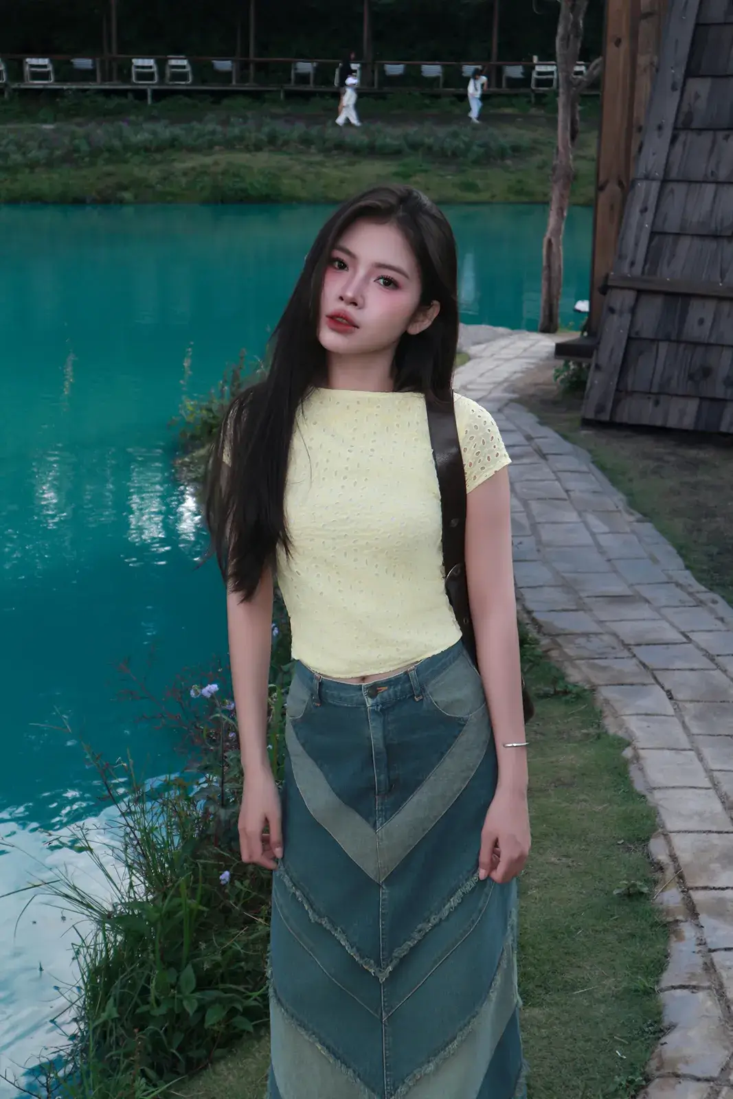 Cô gái mặc áo vàng nhạt và chân váy denim đứng bên hồ nước màu xanh ngọc, phía sau là lối đi lát đá và hàng ghế