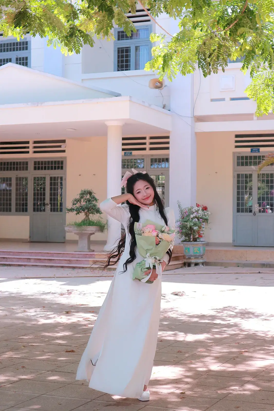 Cô gái mặc áo dài trắng cầm bó hoa đứng trong sân trường, phía sau là dãy phòng học và cây xanh
