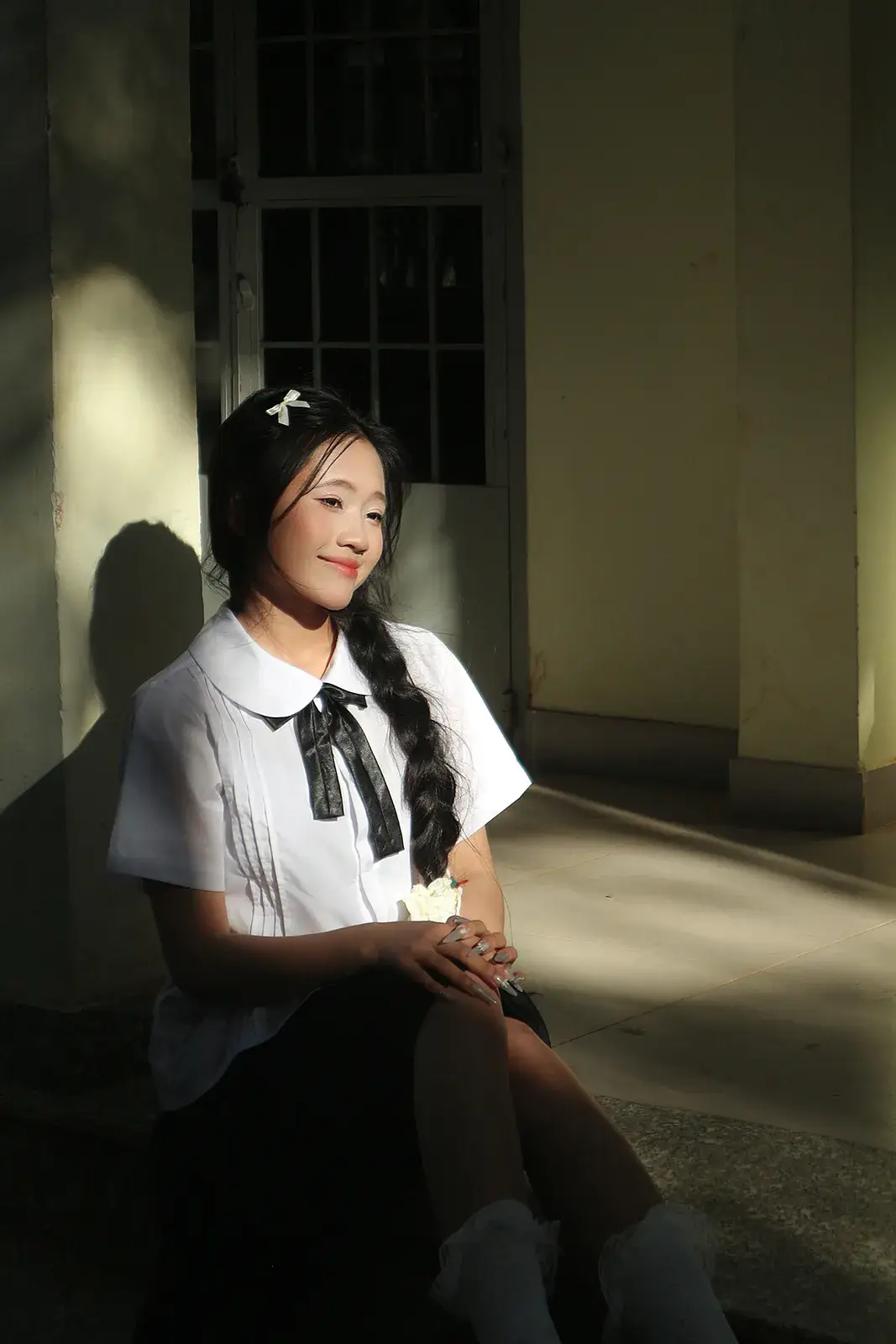 Cô gái mặc áo sơ mi trắng thắt nơ đen, tóc tết, ngồi tựa tường trong vệt nắng, phía sau là khung cửa sổ