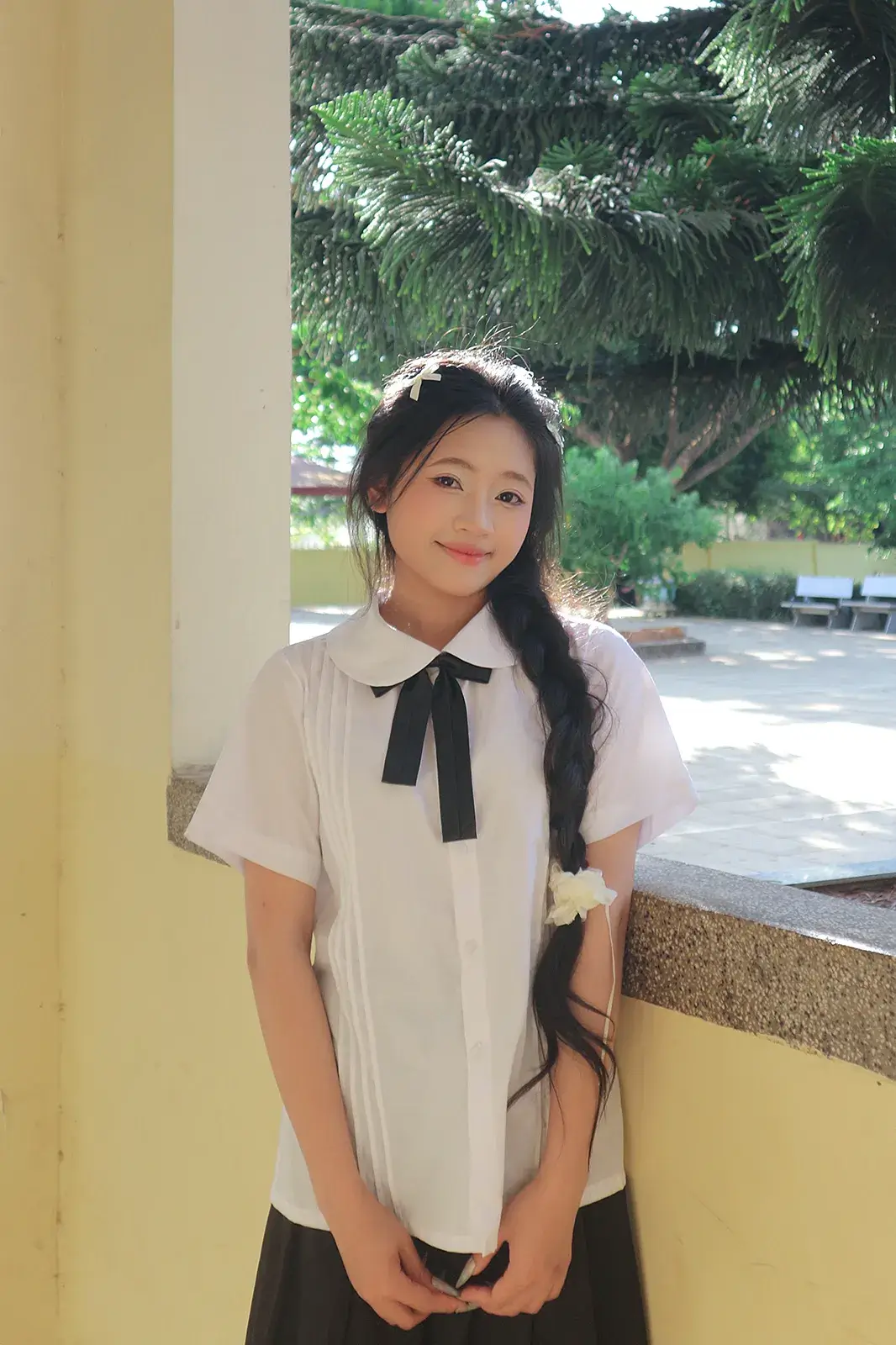 Cô gái mặc áo sơ mi trắng thắt nơ đen, tóc tết, đứng dựa cột hành lang, phía sau là hàng thông và khoảng sân