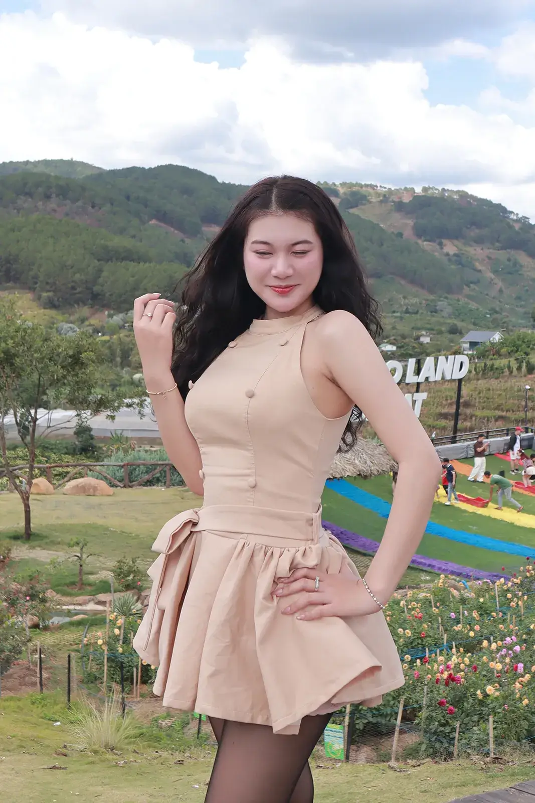 Cô gái mặc váy ngắn màu be đứng trên đồi cỏ, phía sau là rừng thông, vườn hoa và một công trình sọc nhiều màu