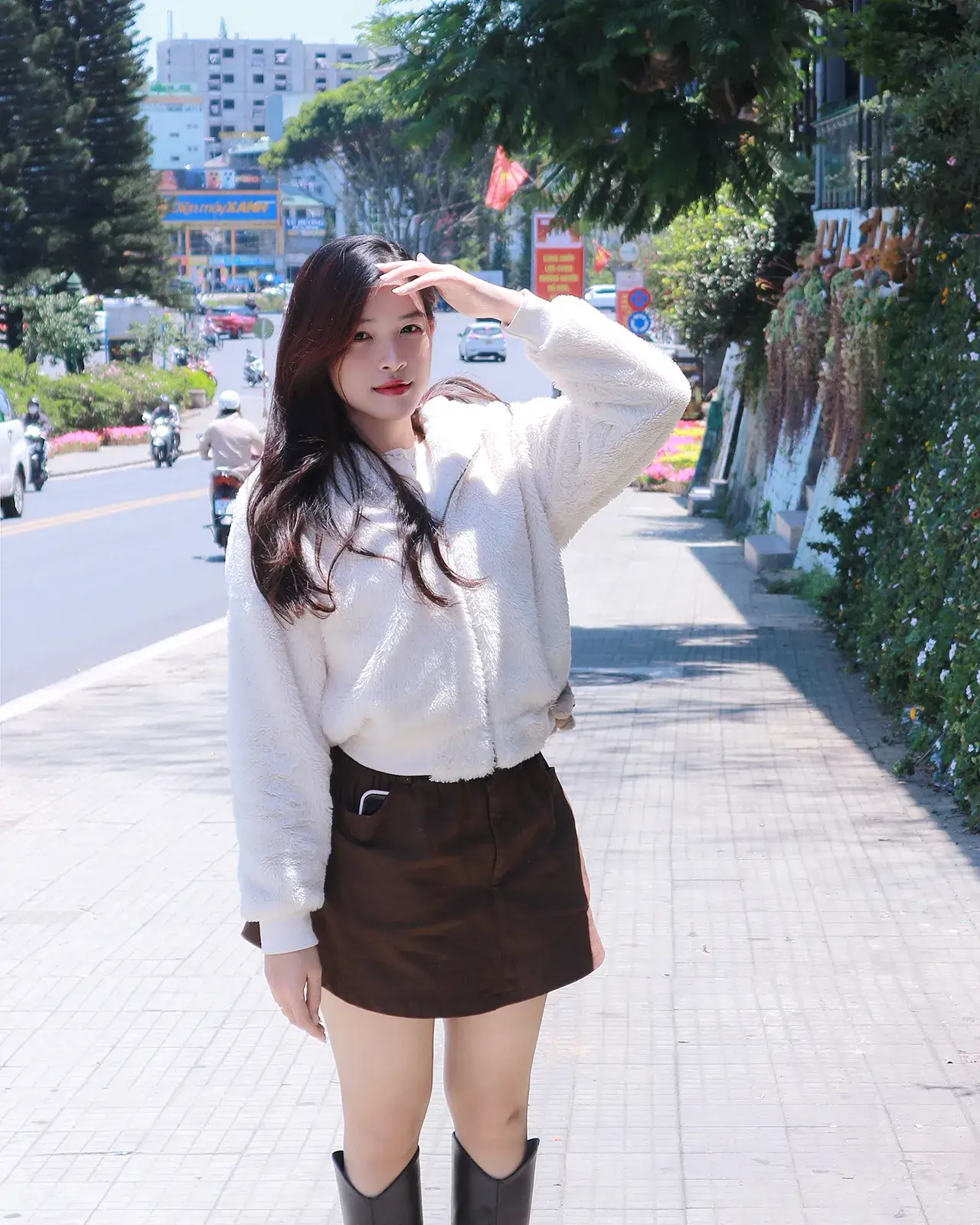 Cô gái mặc áo len trắng, chân váy nâu và bốt cao cổ đứng trên vỉa hè, phía sau là đường phố có xe máy và dãy nhà