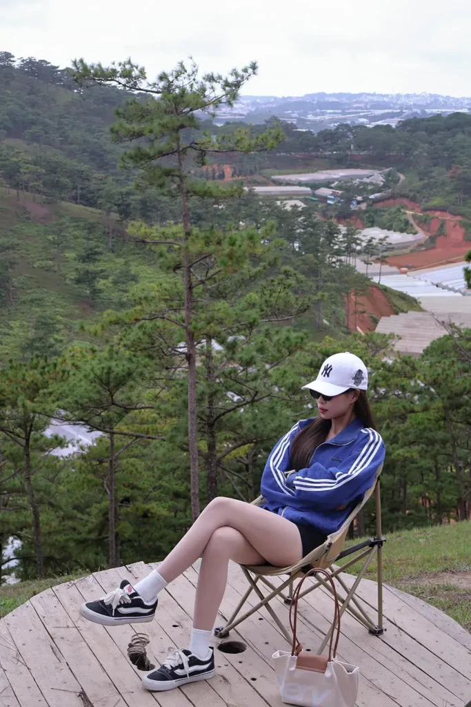 Cô gái mặc áo khoác thể thao xanh ngồi trên ghế xếp trên đồi, nhìn xuống thung lũng thông ở Đà Lạt