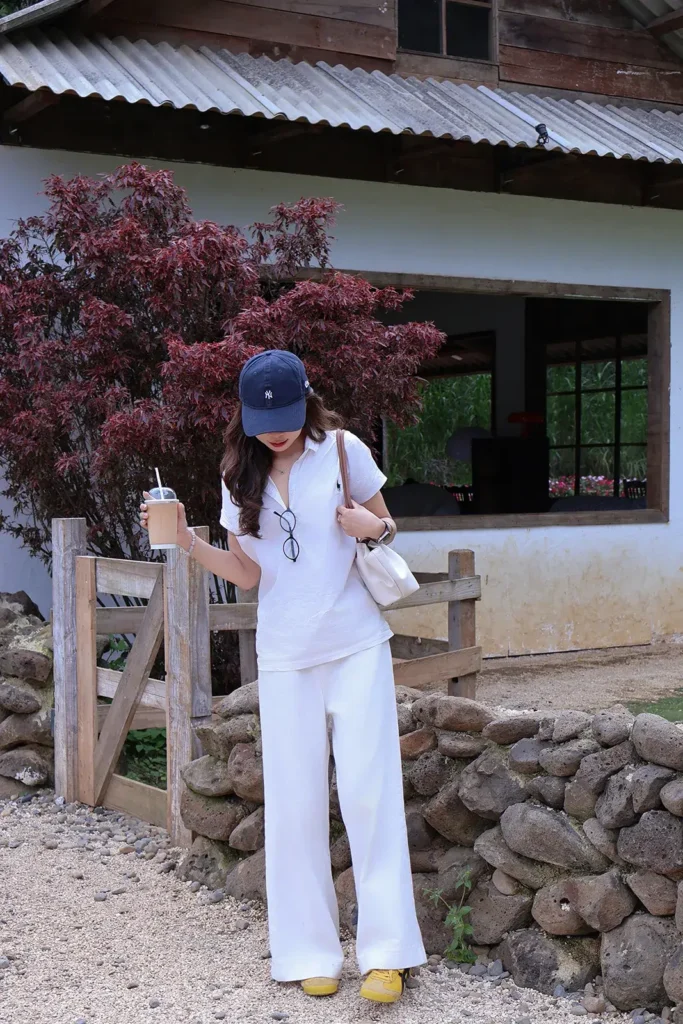 Cô gái mặc đồ trắng đội mũ lưỡi trai cầm ly nước đứng bên hàng rào gỗ trước căn nhà mái tôn ở Đà Lạt