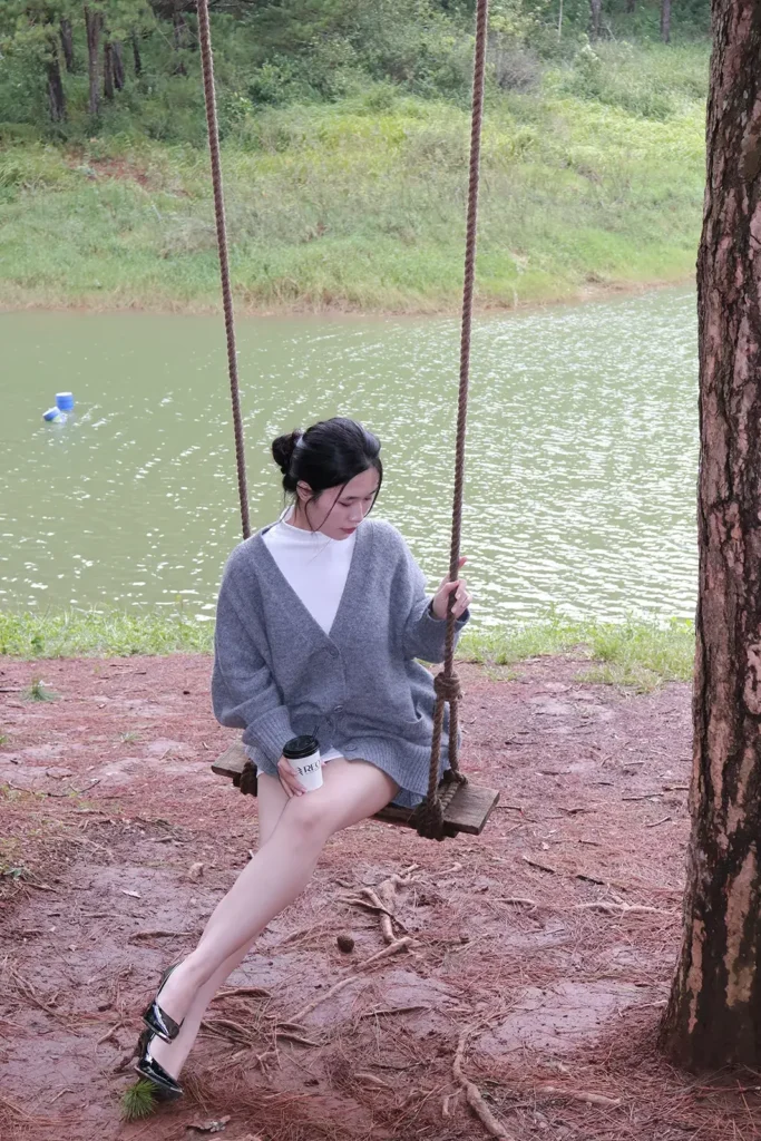Cô gái mặc áo len xám ngồi trên xích đu dây thừng dưới tán thông bên bờ hồ ở Đà Lạt