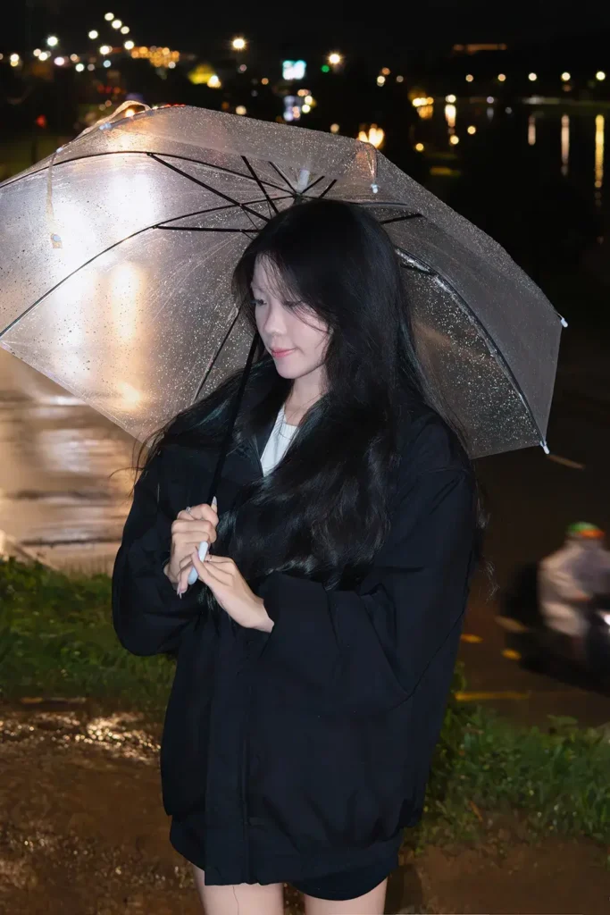 Cô gái mặc áo khoác đen cầm ô đứng ngoài trời buổi tối, phía xa là ánh đèn thành phố Đà Lạt