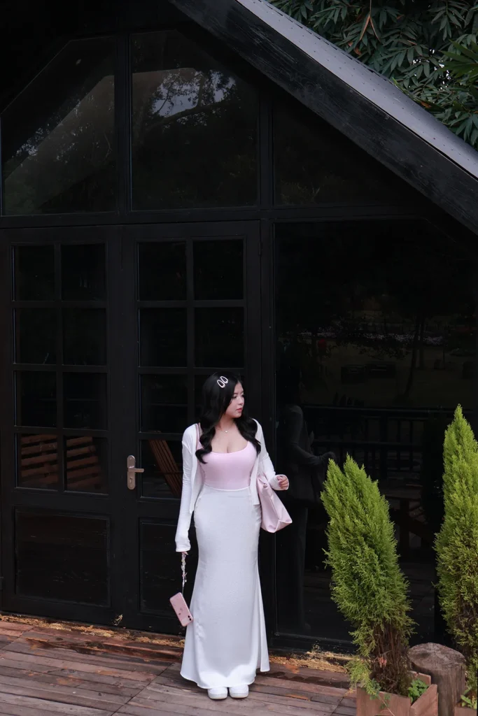 Cô gái mặc váy trắng dài đứng trước căn nhà gỗ sơn đen có cửa kính lớn giữa vườn cây ở Đà Lạt