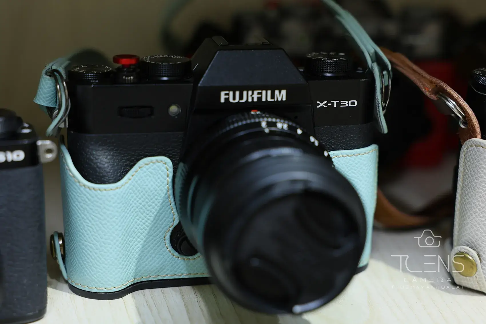 Thân máy Fujifilm X-T30 màu đen gắn ống kính, bọc half-case xanh bạc hà, đặt trên bàn