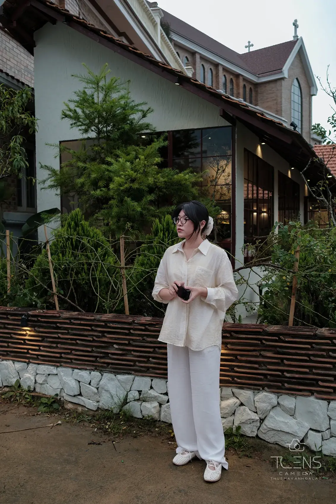 Ảnh chân dung ngoài trời của một phụ nữ mặc đồ trắng đứng bên tường thấp, chụp bằng Fujifilm X-S10 và XF23mm F2