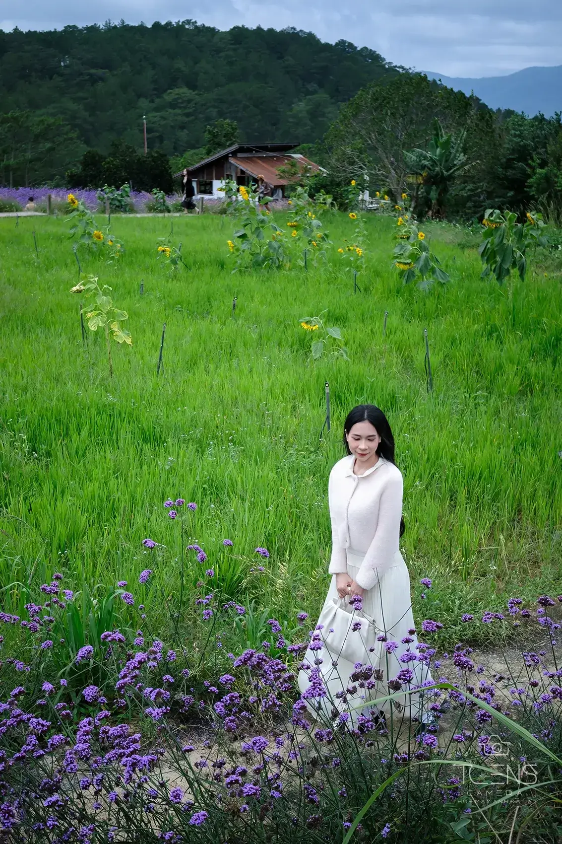 Một phụ nữ mặc váy trắng đứng giữa đồng cỏ có nhà gỗ phía sau, chụp bằng Fujifilm X-T30 với lens 27mm