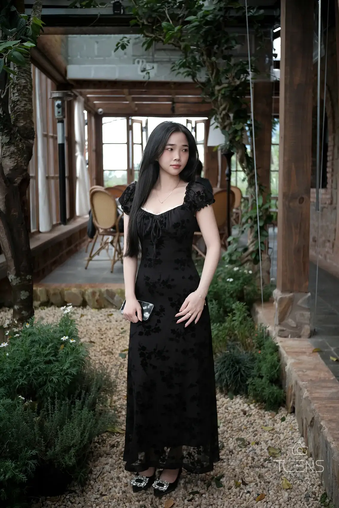 Một phụ nữ mặc váy đen đứng trong lối đi nhà gỗ nhiều cây, chụp bằng Fujifilm X-T30 với lens 27mm